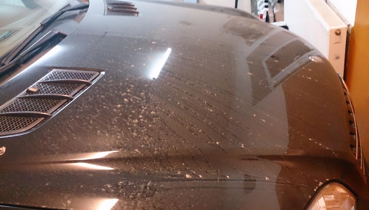 Brudna, czarna maska samochodu z widocznymi smugami i osadem, sugerująca potrzebę umycia. Widoczny fragment reflektora i kratki wentylacyjnej.