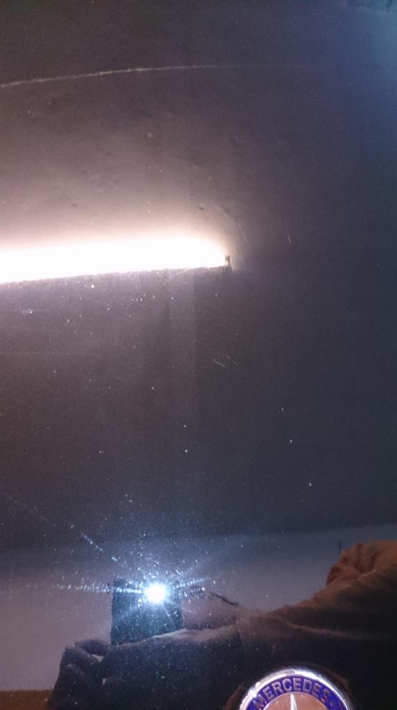 Błyszcząca, ciemna powierzchnia z widocznymi smugami i odbiciami światła, prawdopodobnie karoseria samochodu po polerowaniu, z fragmentem emblematu Mercedes-Benz.