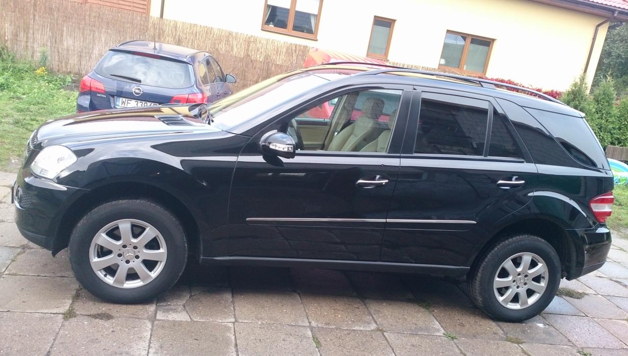 Czarny Mercedes ML na kostce brukowej, widok z boku, z innym autem w tle. Oświetlenie dzienne, dom w tle. Felgi aluminiowe.