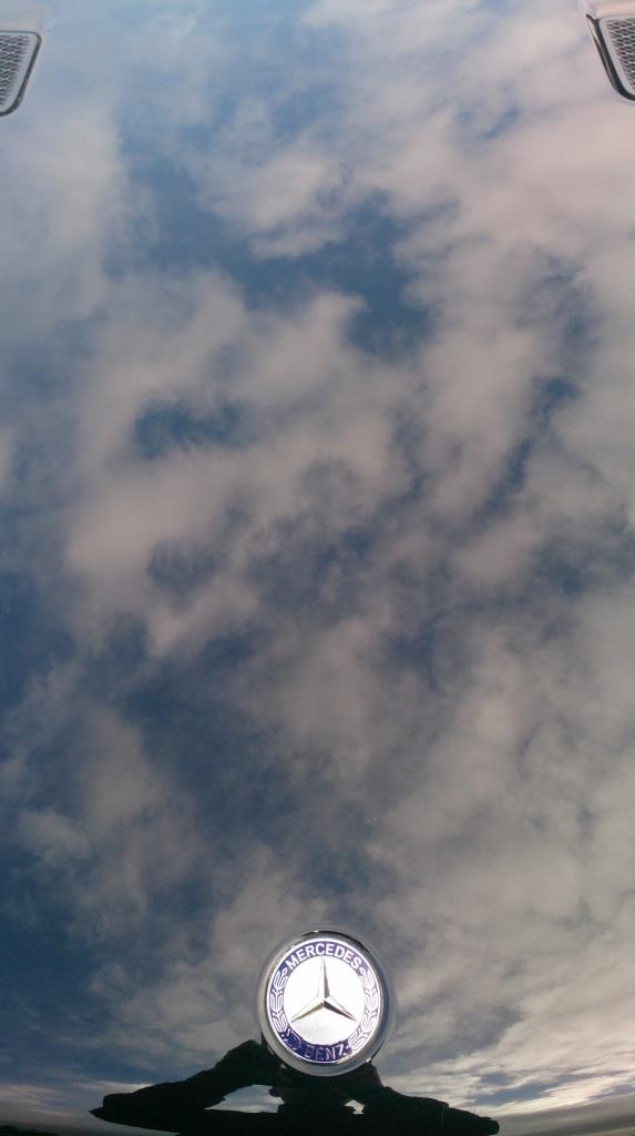 Błyszcząca maska czarnego Mercedesa odbija błękitne niebo z chmurami. Widoczne logo marki. Kąt ujęcia z dołu podkreśla głębię.