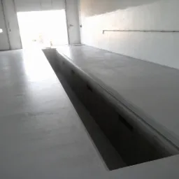 Szara, gładka wylewka w hali z kanałem rewizyjnym. Oświetlenie dzienne wpada przez otwarte wrota garażowe. Widoczna instalacja elektryczna na ścianie.