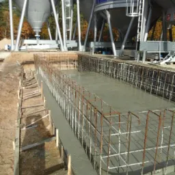 Zalewanie fundamentów pod konstrukcję przemysłową. Widoczne szalunki, zbrojenie i silosy w tle. Etap wylewki.