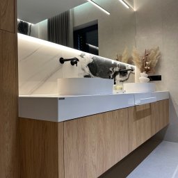 Mexbud - Nowoczesna łazienka z dwoma białymi umywalkami nablatowymi, drewnianą szafką pod umywalką i podświetlanym lustrem. Minimalistyczny design z elementami dekoracyjnymi.