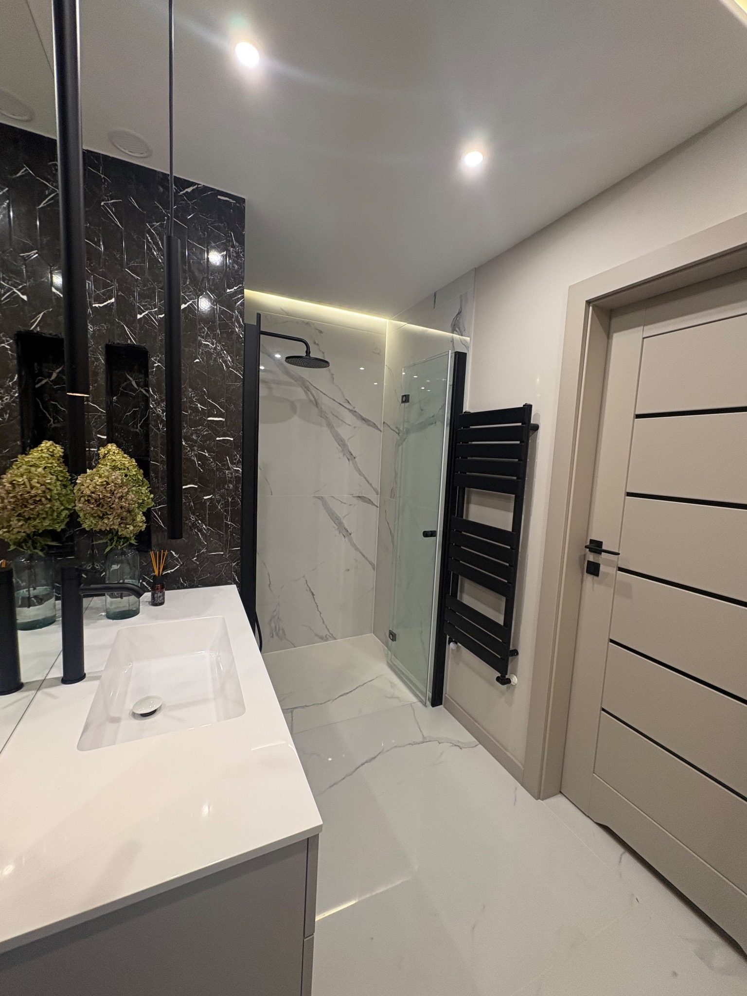 Nowoczesna łazienka z kabiną prysznicową typu walk-in, umywalką nablatową i czarnym grzejnikiem drabinkowym. Marmurowe płytki na ścianach i podłodze.