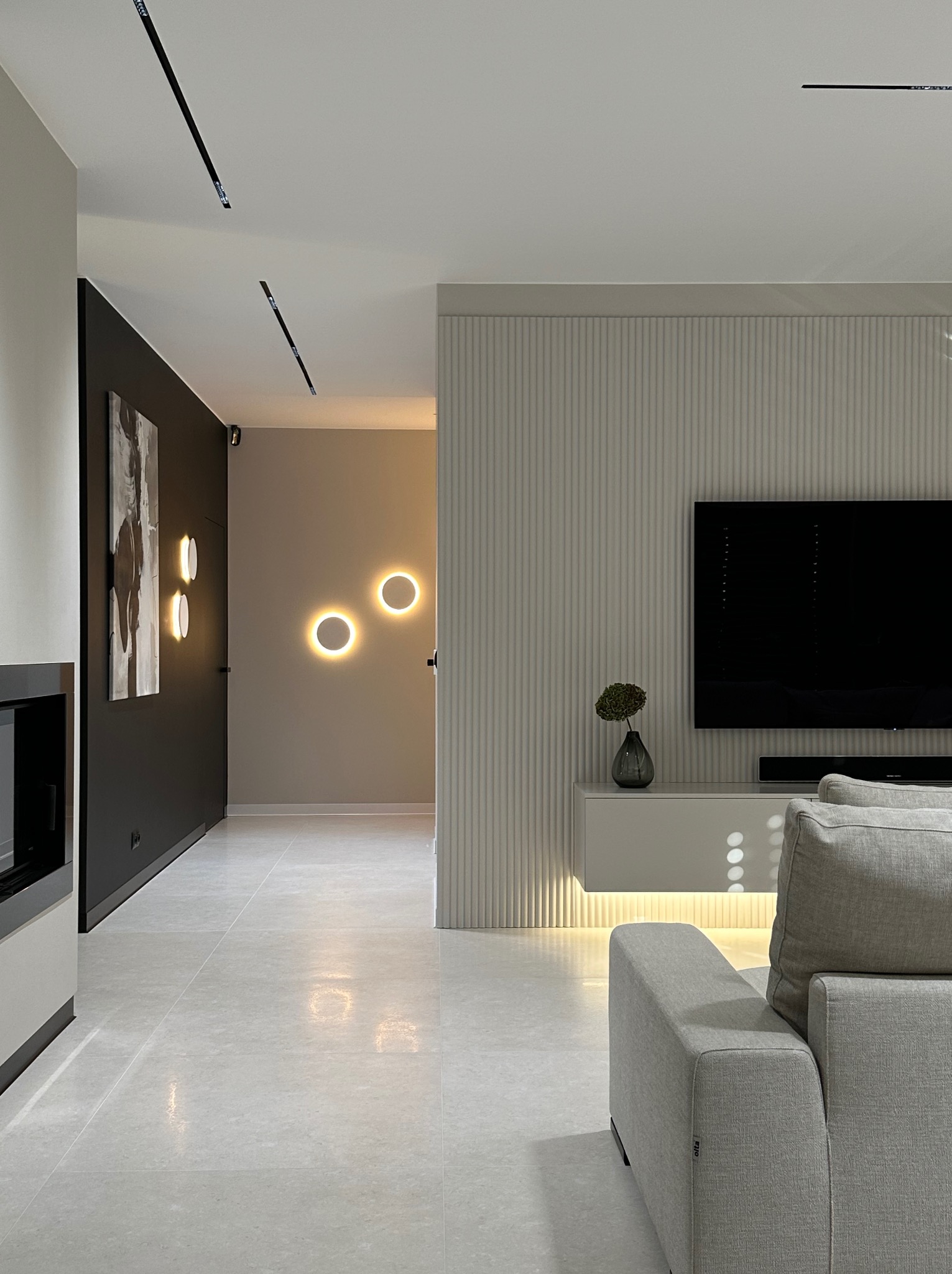 Nowoczesne wykończenie wnętrza: szara sofa, telewizor na ścianie z lamelami, oświetlenie punktowe i minimalistyczny design. Widok na korytarz z okrągłymi lampami.