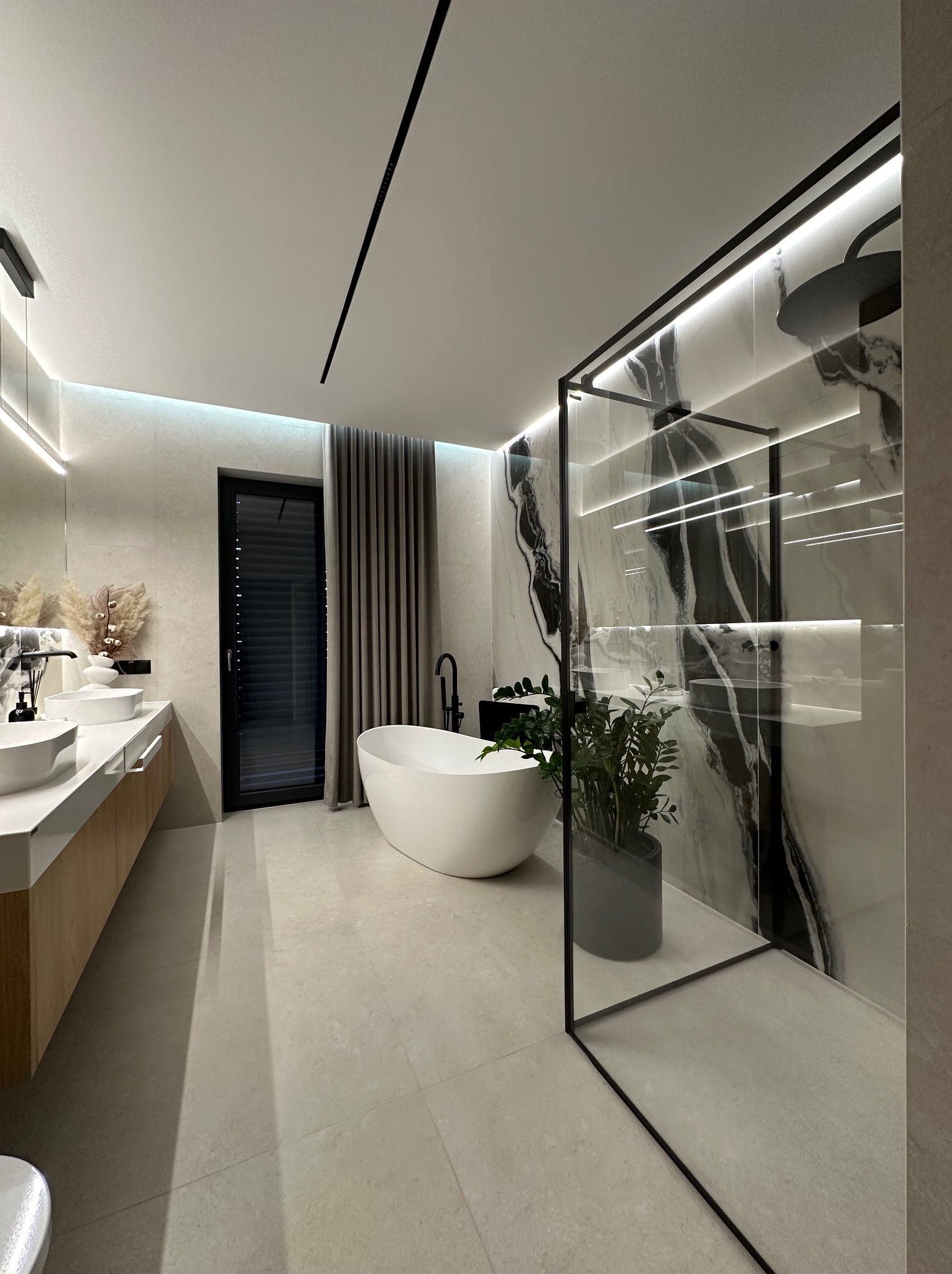 Nowoczesna łazienka z wolnostojącą wanną, kabiną prysznicową typu walk-in z marmurowym wykończeniem i drewnianą szafką pod umywalką. Minimalistyczne oświetlenie.