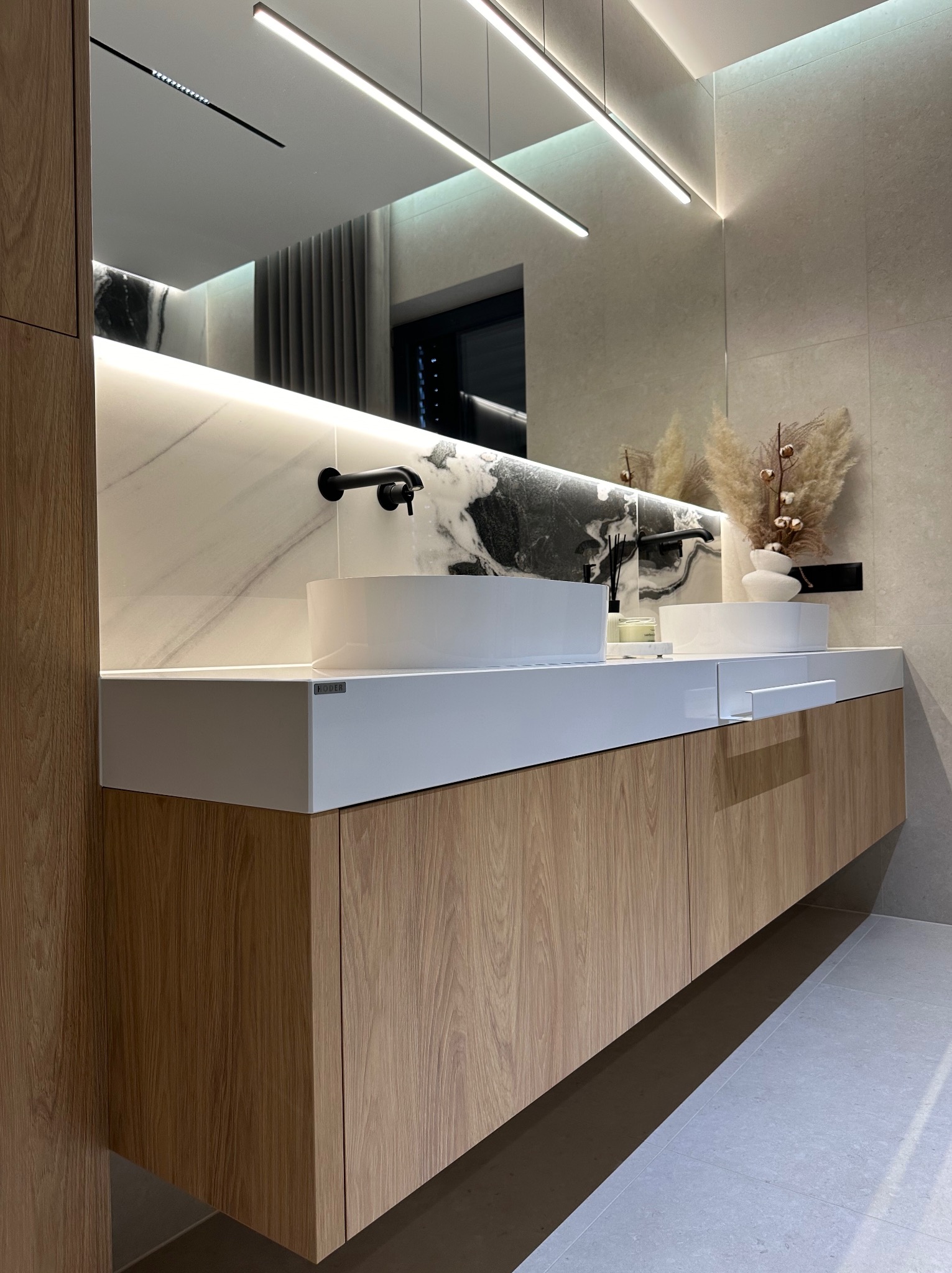 Nowoczesna łazienka z dwoma białymi umywalkami nablatowymi, drewnianą szafką pod umywalką i podświetlanym lustrem. Minimalistyczny design z elementami dekoracyjnymi.