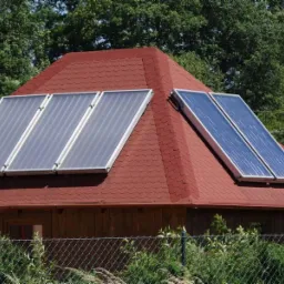 Czerwony dach z panelami solarnymi, widoczne zza ogrodzenia. Trzy panele po lewej i dwa panele po prawej stronie dachu. W tle drzewa.