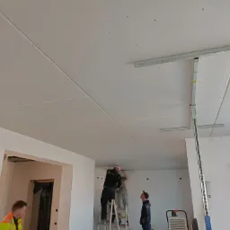 Montaż sufitu podwieszanego z płyt kartonowo-gipsowych: dwóch monterów na drabinie przytwierdza płyty do metalowej konstrukcji, trzeci obserwuje, widoczne profile, śruby i poziomica.