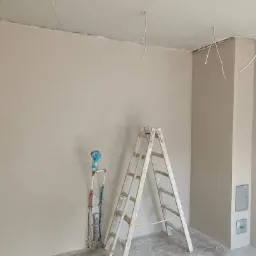 Wnętrze po nałożeniu gładzi szpachlowej na ścianach i suficie, widoczne wystające kable elektryczne, drabina malarska i instalacja wodna zabezpieczona folią.