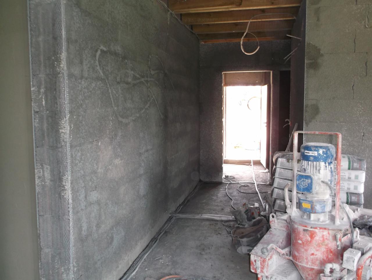 Wnętrze w trakcie tynkowania maszynowego: ściany pokryte szarym tynkiem, widoczna maszyna do tynkowania i worki z zaprawą. Metalowe narożniki zamontowane.