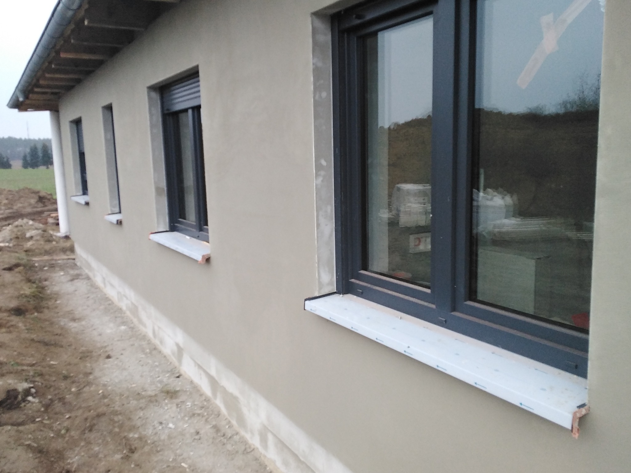 Świeżo otynkowana ściana zewnętrzna domu jednorodzinnego z szarymi oknami i roletami, widok z boku na tle krajobrazu polnego.