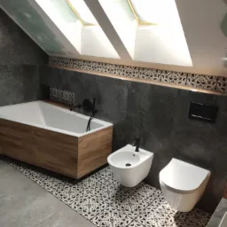 Nowoczesna łazienka na poddaszu z drewnianą obudową wanny, bidetem i toaletą podwieszaną, wykończona szarą płytką i dekoracyjnym pasem płytek nad wanną, widoczne okno dachowe.