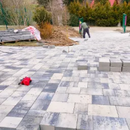 Pracownik w trakcie układania dwukolorowej kostki brukowej na podwórku, widoczne ułożone fragmenty nawierzchni, paleta z kostką, czerwone rękawice robocze i narzędzia.