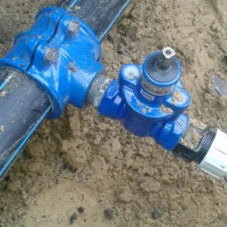 Niebieski zawór odcinający na czarnym rurociągu z niebieskim paskiem, częściowo zakopany w piasku.