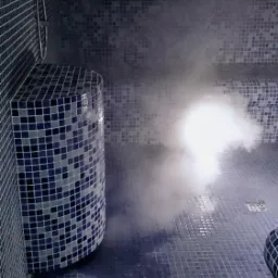 Wnętrze łaźni parowej wyłożone niebieską mozaiką, z widoczną dyszą pary i siedziskiem w rogu.