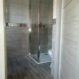 Wykończona łazienka z szarymi płytkami imitującymi drewno na podłodze, kabiną prysznicową z chromowanymi profilami i dekoracyjnym pasem mozaiki nad płytkami ściennymi. Częściowo widoczna toaleta.