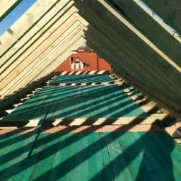 Wnętrze konstrukcji drewnianego dachu w trakcie budowy, widok z dołu na zieloną membranę dachową i krokwie, w tle czerwony dach sąsiedniego budynku.