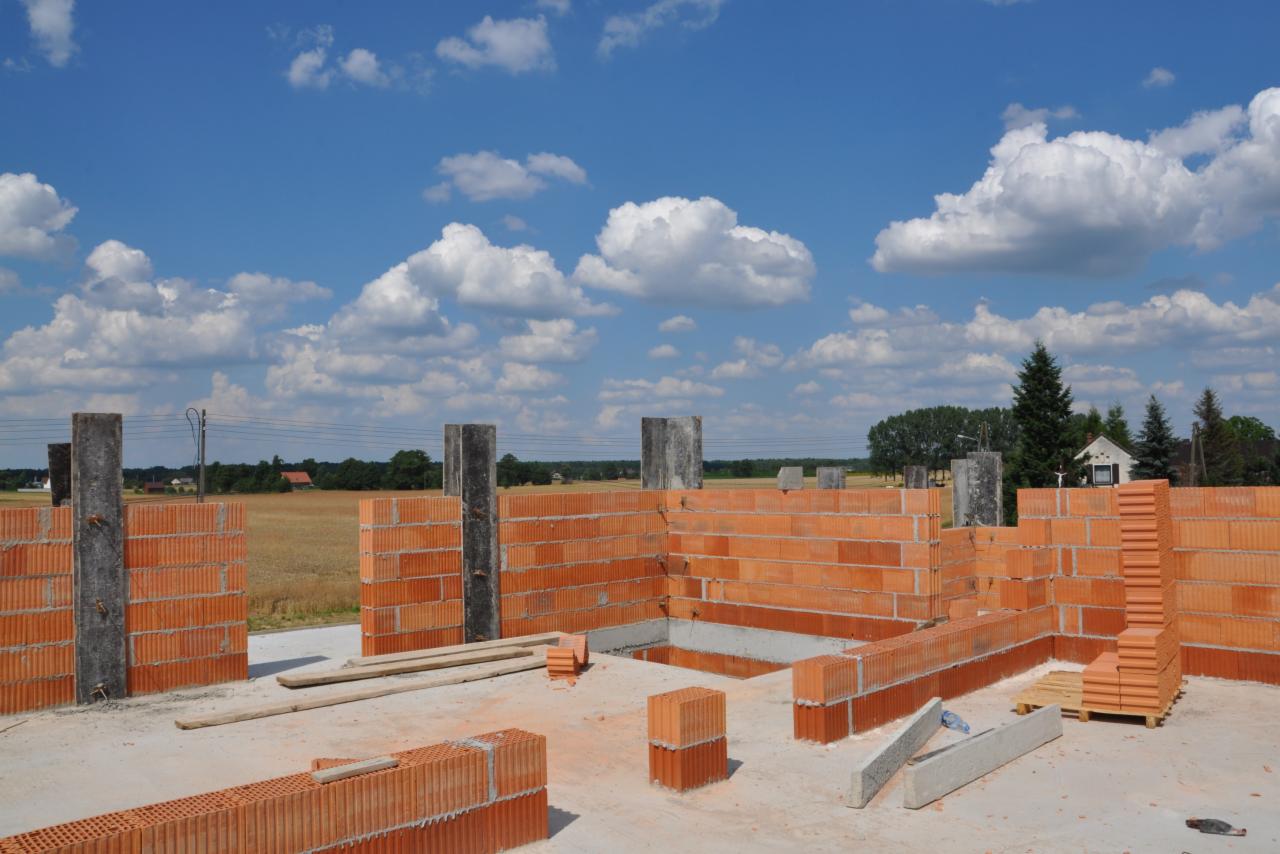 Wznoszenie ścian domu z czerwonej cegły ceramicznej. Widoczne betonowe słupy, fragment fundamentów, w tle pola i błękitne niebo z chmurami.