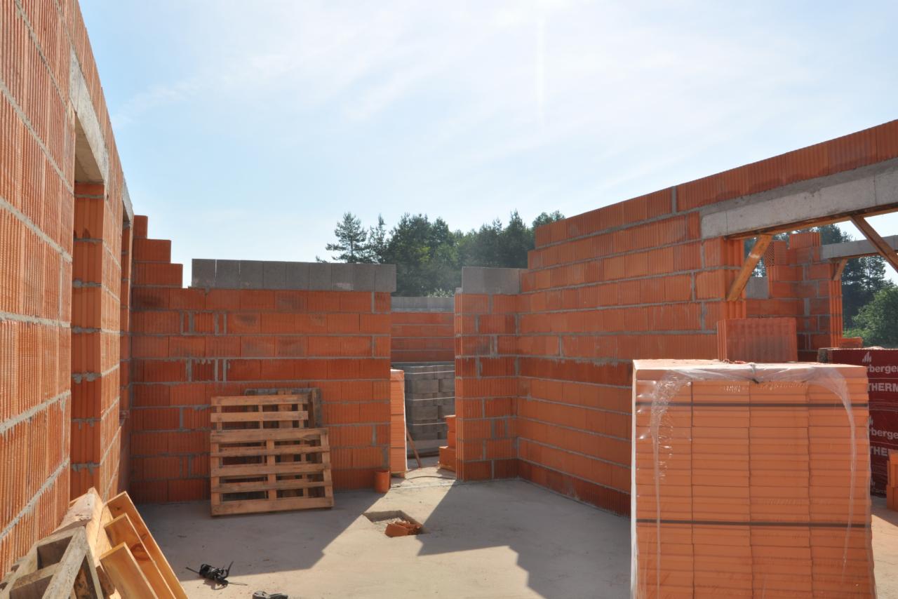 Wnętrze budowanego domu z czerwonej cegły. Widoczne palety i zapakowane cegły. W tle zieleń drzew i błękit nieba. Surowy stan budynku.