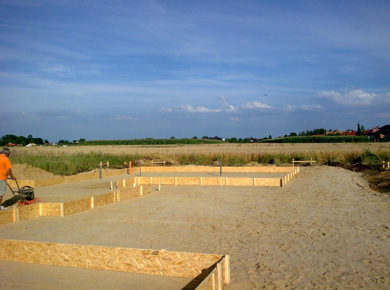 Wykop pod fundamenty domu z drewnianym szalunkiem, zagęszczany przez pracownika. W tle pole i zabudowania pod błękitnym niebem.