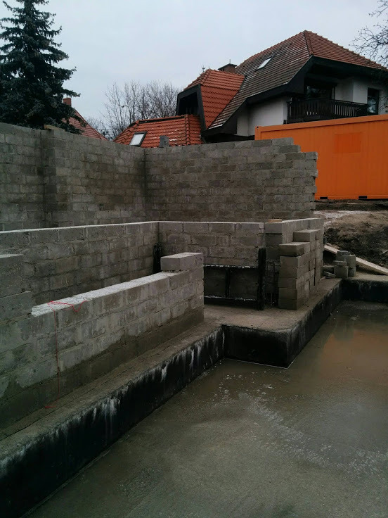 Wylany fundament z częściowo wzniesionymi ścianami z bloczków betonowych, widoczna izolacja fundamentu, w tle budynek mieszkalny z czerwoną dachówką i pomarańczowy kontener budowlany.