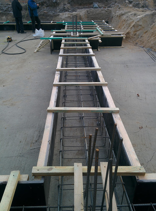 Szczegółowe ujęcie z góry na szalunki fundamentowe zbrojone stalą, z widocznymi elementami drewnianymi i stalowymi prętami, przygotowane do zalania betonem, na tle ziemi i dwóch osób w oddali.