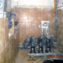 Pomieszczenie techniczne z trzema pompami Grundfos na metalowej rurze, brązowe płytki na ścianach i podłodze, instalacja wodna. Widoczne elementy instalacji i narzędzia.