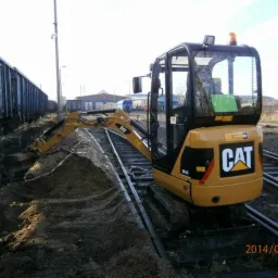 Żółta minikoparka CAT na torach kolejowych, wykonująca wykop w ziemi, obok stoi wagon towarowy.