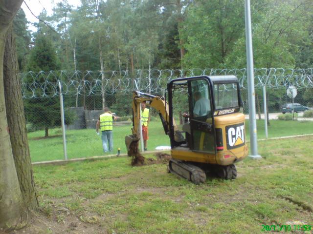 Żółta minikoparka CAT podczas pracy przy ogrodzeniu z drutem kolczastym; w tle widoczni pracownicy w kamizelkach odblaskowych i zalesiony teren.