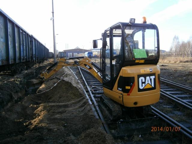 Żółta minikoparka CAT na torach kolejowych, wykonująca wykop w ziemi, obok stoi wagon towarowy.