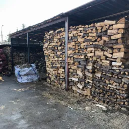 Zadaszony skład z ułożonymi rzędami pociętego drewna, obok biały samochód dostawczy i duży worek z drewnem w tle.