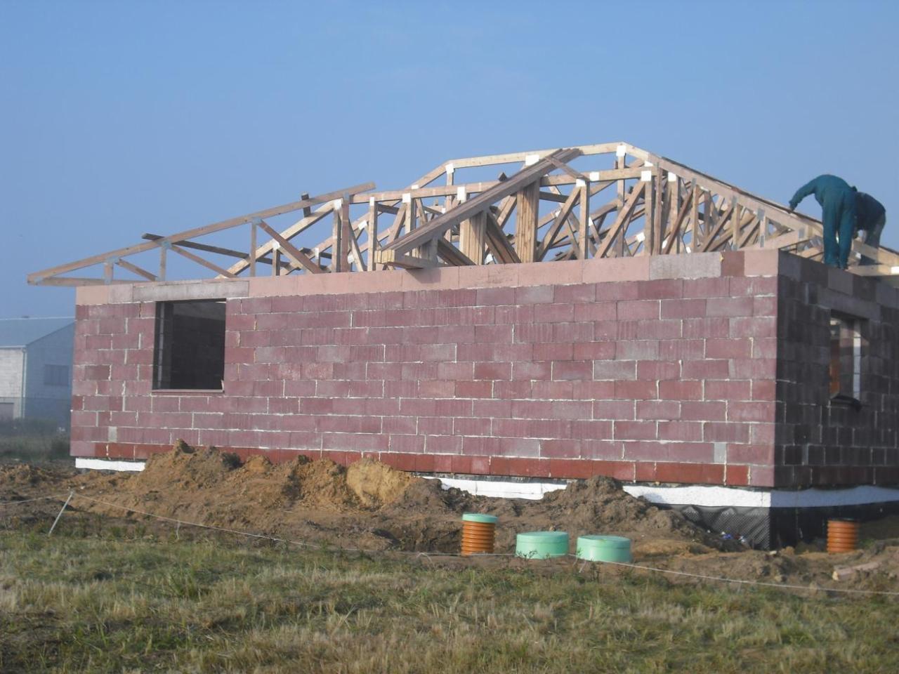 Wznoszenie ścian domu z bloczków keramzytobetonowych, widoczna konstrukcja dachu, dwóch robotników na górze, fundament zabezpieczony folią, w tle trawa i budynek.