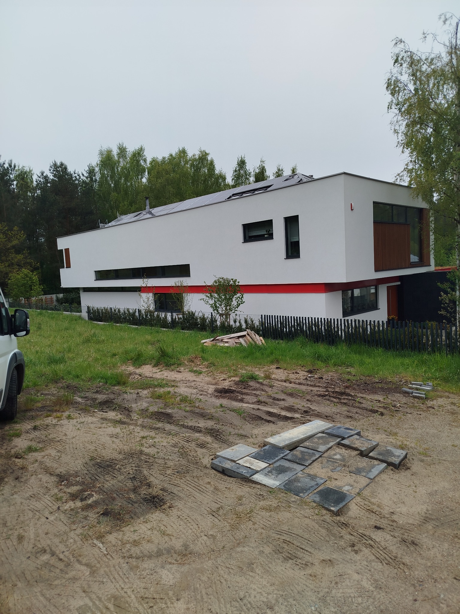 Nowoczesny, biały dom z płaskim dachem i panelami słonecznymi, z czerwoną linią akcentującą, otoczony zielenią i częściowo ułożoną kostką brukową.