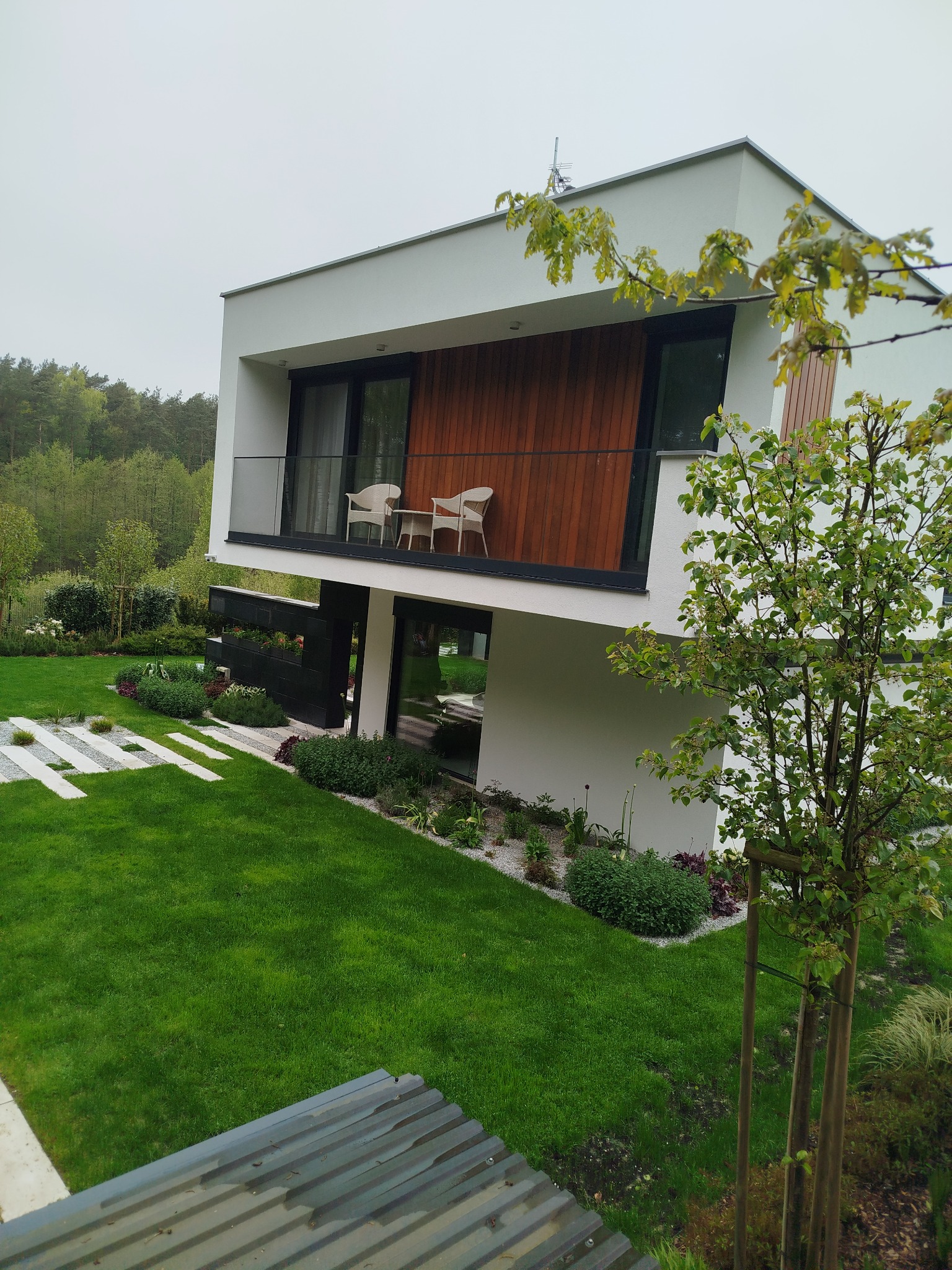 Nowoczesny dom z drewnianą elewacją balkonu, widok z góry na zielony trawnik i ścieżkę z płyt. Minimalistyczny design, naturalne otoczenie.