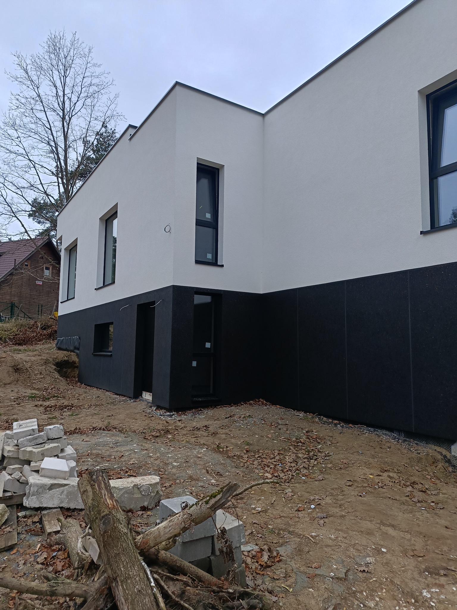 Nowoczesny dom z białą elewacją i czarnym cokołem, widoczny z zewnątrz, z elementami krajobrazu i stertą materiałów budowlanych w pobliżu.