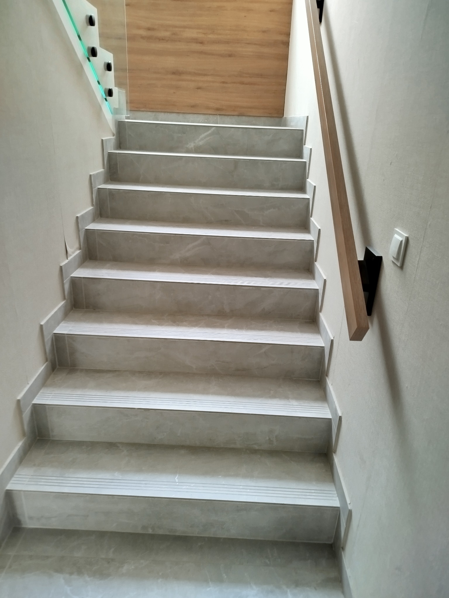Wykończone schody wewnętrzne z szarymi płytkami imitującymi marmur, z drewnianą poręczą i szklaną balustradą. Widok z dołu na górę.