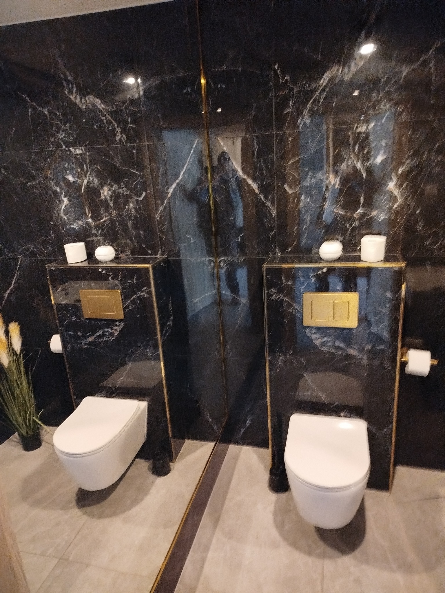 Elegancka, wykończona łazienka z czarnymi marmurowymi płytkami, podwieszaną toaletą i złotymi akcentami. Widoczne detale wykończenia i oświetlenie.