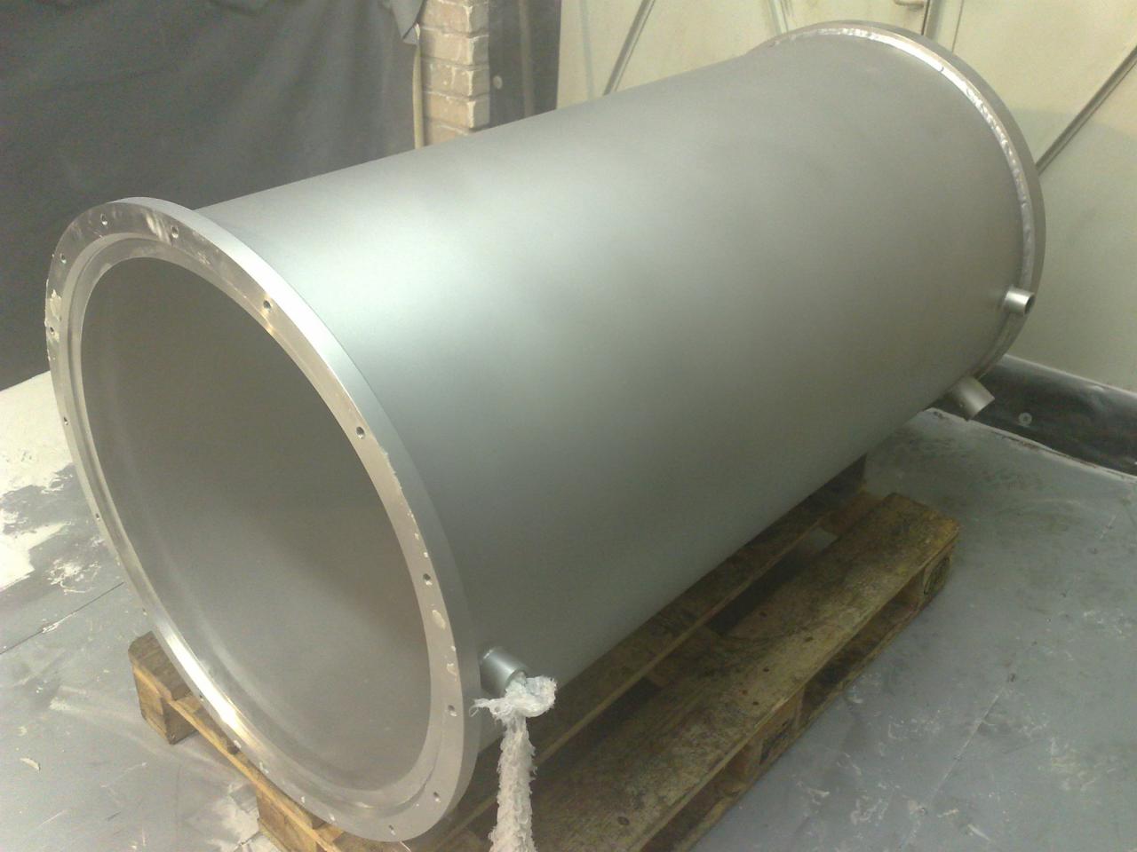 Szary, cylindryczny element metalowy z kołnierzem na palecie, prawdopodobnie po malowaniu proszkowym. Widoczne otwory i spawane elementy.