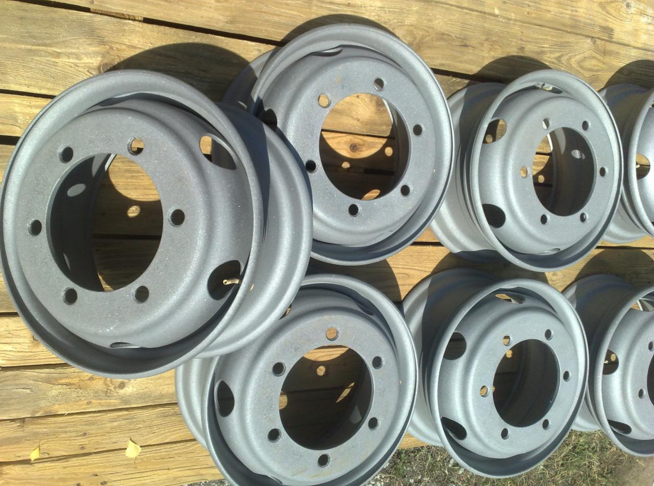 Szare, metalowe felgi samochodowe z otworami, ułożone na drewnianym podłożu. Widoczne cienie i faktura powierzchni felg. Ujęcie z góry, ostre światło.