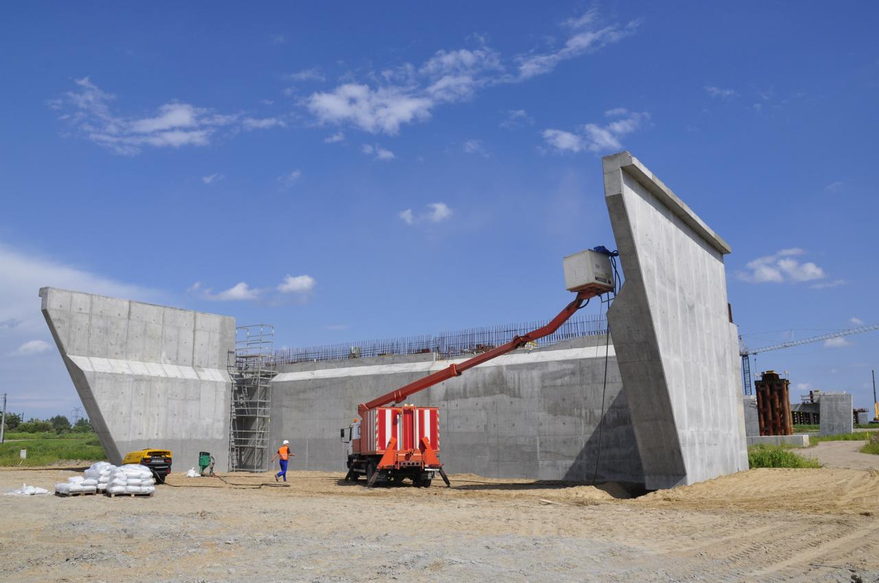 Budowa betonowego mostu w toku: widoczny fragment konstrukcji z użyciem podnośnika koszowego, rusztowania i pracownika w odblaskowej kamizelce na placu budowy.