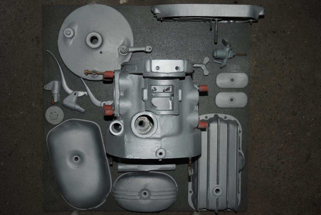 Elementy silnika motocyklowego po piaskowaniu i malowaniu, rozłożone na szarym tle. Widoczne detale, takie jak dźwignie, pokrywy i korpus silnika.