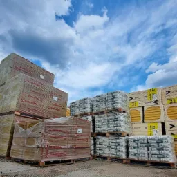 Sztaple materiałów budowlanych: wełna mineralna Rockwool, styropian TermPIR XL oraz worki z zaprawą Bolix, ułożone na paletach pod błękitnym niebem z chmurami.