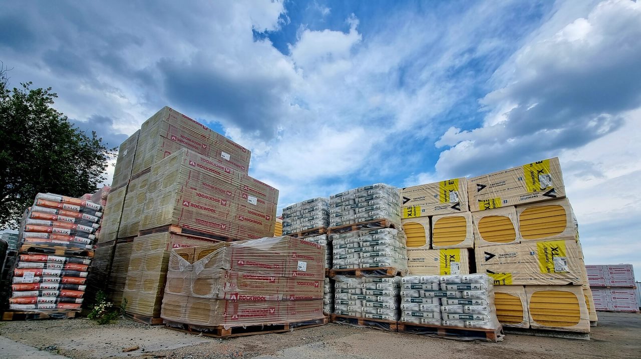 Sztaple materiałów budowlanych: wełna mineralna Rockwool, styropian TermPIR XL oraz worki z zaprawą Bolix, ułożone na paletach pod błękitnym niebem z chmurami.