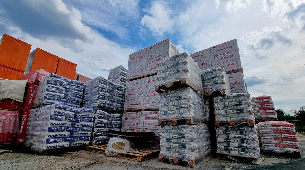 Sztaple materiałów budowlanych różnych producentów, w tym worki Kreisel i płyty Rockwool, ułożone na paletach pod błękitnym niebem.