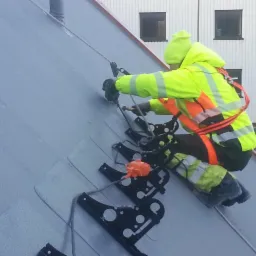 Pracownik w odzieży ochronnej, zabezpieczony linami, montuje elementy na szarej, ukośnej powierzchni, prawdopodobnie dachu, z wykorzystaniem specjalistycznego sprzętu i czarnych uchwytów z białymi...