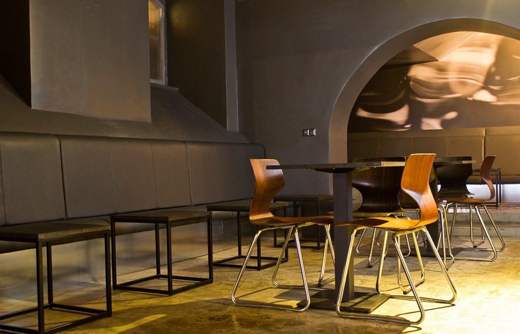 Wnętrze lokalu komercyjnego z szarymi ścianami, długą tapicerowaną ławą, małymi stolikami i krzesłami z giętego drewna na metalowych nogach, oświetlone ciepłym światłem