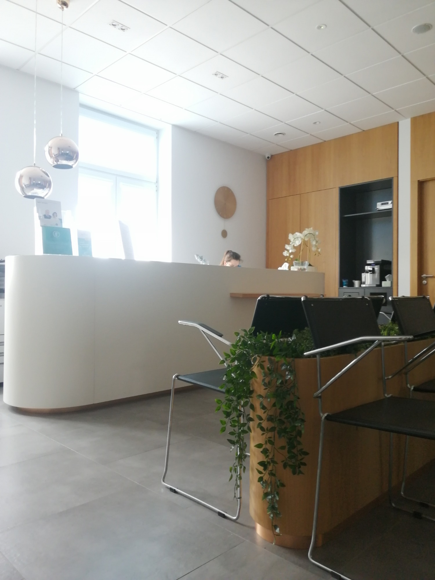 Jasne wnętrze przychodni z recepcją o zaokrąglonym kształcie, nowoczesne krzesła z roślinami doniczkowymi, minimalistyczny wystrój z drewnianymi elementami i okrągłym zegarem na ścianie.