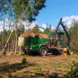 Zielony kombajn leśny wypełnia przyczepę zrębkami drzewnymi na skraju lasu, pracownik w pomarańczowym kombinezonie nadzoruje proces.