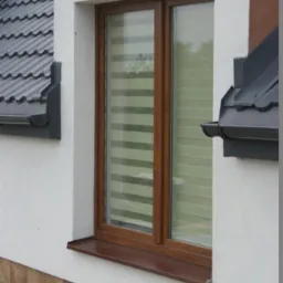 Brązowe okno z roletami dzień-noc w białej elewacji budynku. Widoczny fragment dachu z rynnami. Ujęcie z poziomu chodnika z brązowych płytek.